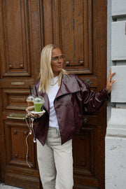 Veste en simili cuir loose portée par une femme, silhouette entière, devant une porte en bois, gobelet à la main
