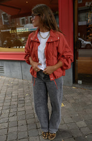 Veste courte déperlante portée par une femme, silhouette entière, t-shirt blanc, jean gris, vitrine rouge