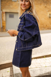 Veste en jean courte portée par une femme, vue de trois-quarts, mur en pierre, jupe en jean