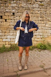 Veste en jean courte portée par une femme, vue de face, mur en pierre, sac en toile