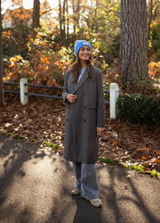 Veste longue boutonnée doublée grise portée par une femme, silhouette entière, bonnet bleu, sac