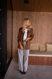 Veste effet daim ceinturée chocolat portée par une femme, silhouette entière, mur en bois, pantalon gris