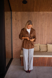 Veste effet daim ceinturée chocolat portée par une femme, silhouette entière, pantalon gris, banc