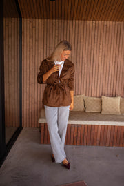 Veste effet daim ceinturée chocolat portée par une femme, silhouette entière, banc, pantalon gris