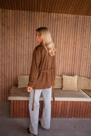 Veste effet daim ceinturée chocolat portée par une femme, vue de dos, mur en bois, pantalon gris