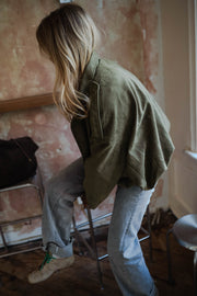 Veste loose à zip doré kaki portée par une femme, vue de profil, tabouret, mur patiné