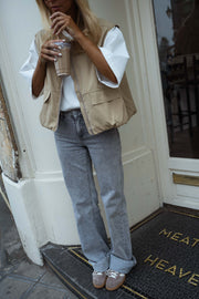 Veste sans manches à poches beige portée par une femme, vue de face, porte vitrée, gobelet