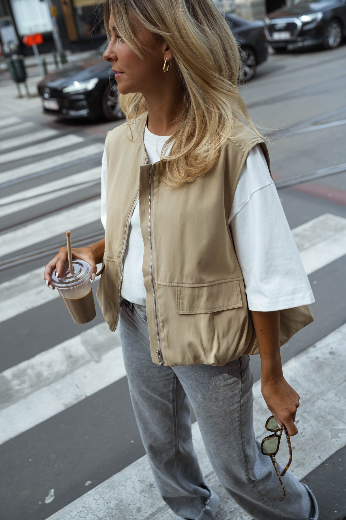 Veste sans manches à poches beige portée par une femme, en marchant, passage piéton, gobelet