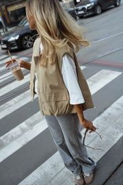 Veste sans manches à poches beige portée par une femme, vue de trois-quarts, passage piéton, gobelet