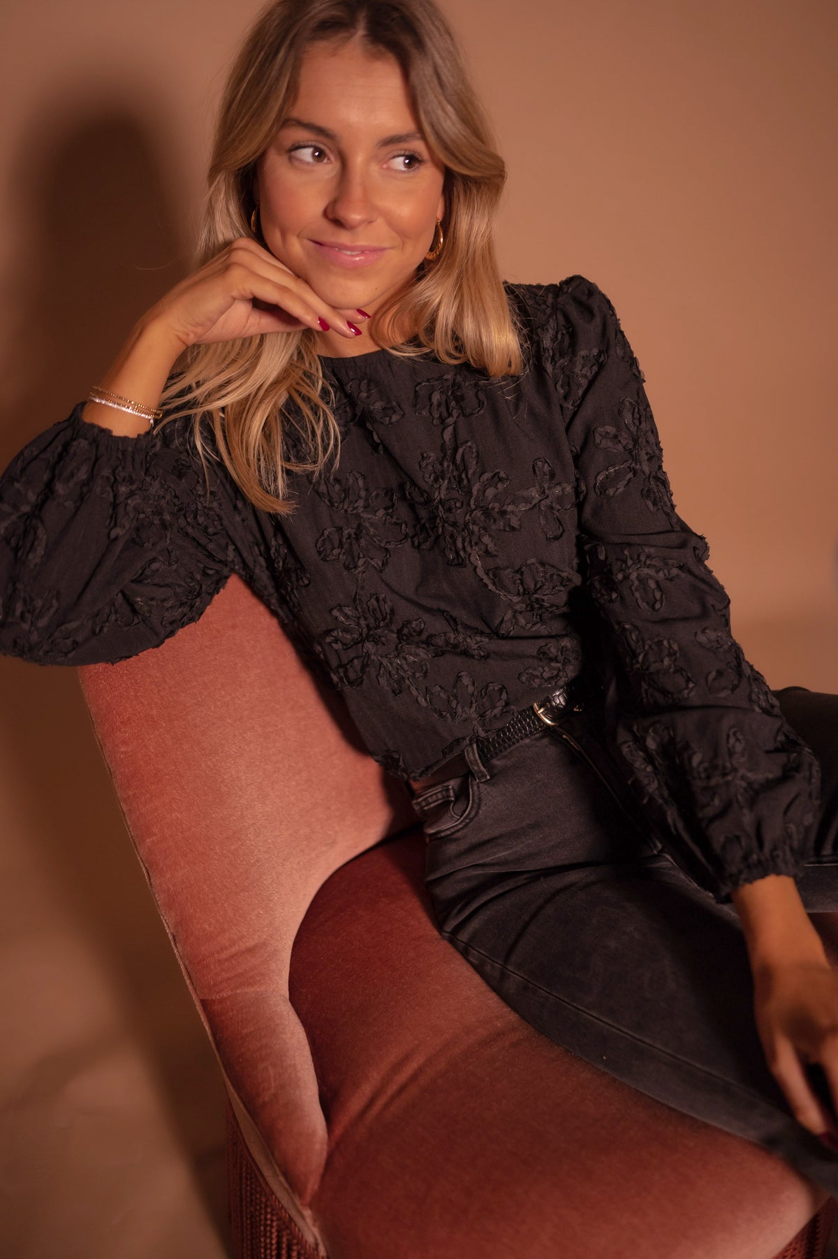 Blouse à poignets élastiques portée par une femme, assise, fauteuil rose, jean noir, main sur le menton