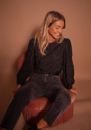 Blouse à poignets élastiques portée par une femme, assise, sur un fauteuil rose, jean noir, de face