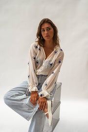 Blouse transparente à fleurs brodées portée par une femme, assise, tabouret, jean large