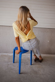 Blouse dos ouvert à nouer jaune portée par une femme, vue de dos, tabouret bleu, mur gris