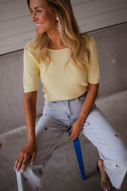 Blouse dos ouvert à nouer jaune portée par une femme, vue de face, tabouret bleu, jean clair