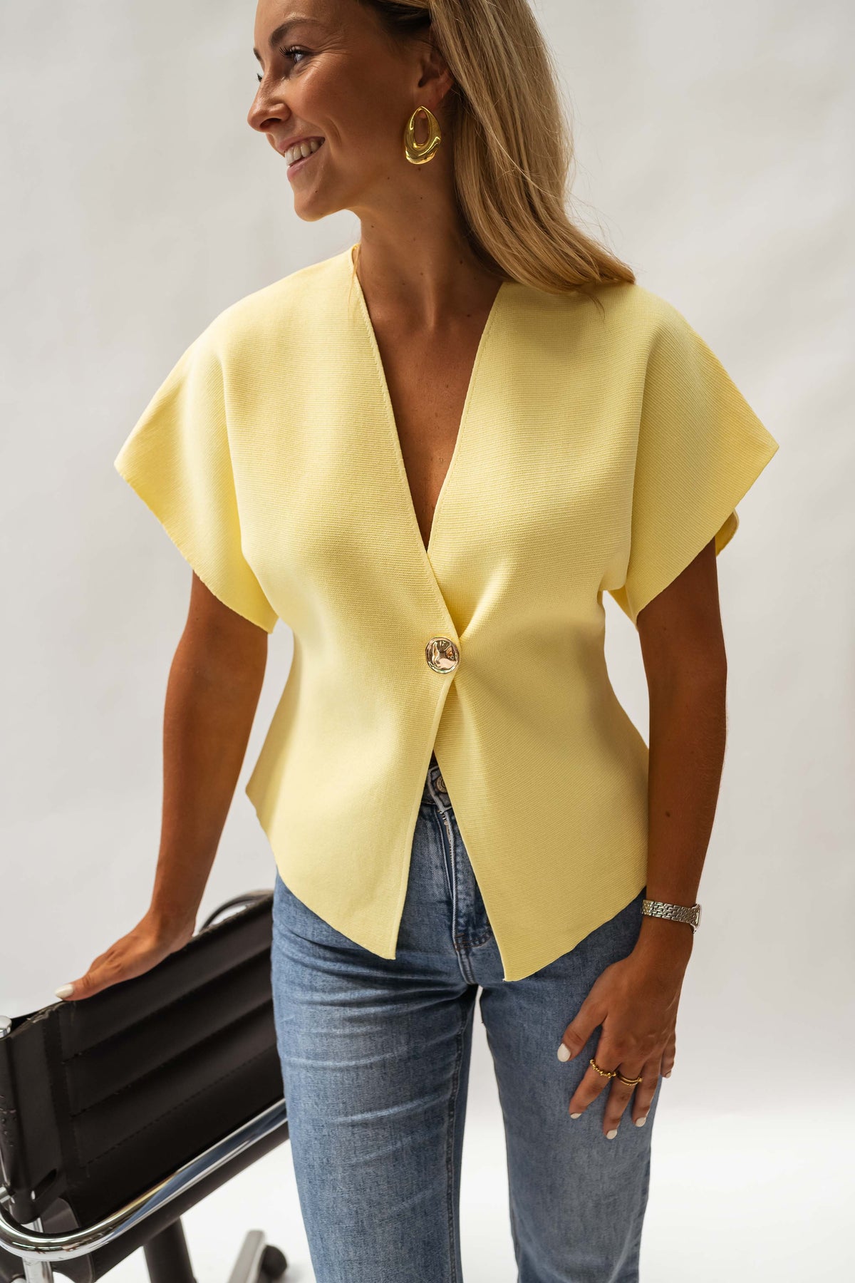Blouse évasée à col V jaune portée par une femme, vue de trois-quarts, bouton, chaise noire