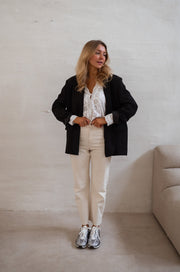Blouse brodée aux poignets portée par une femme, silhouette entière, blazer noir par-dessus, pantalon beige