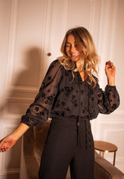 Blouse fleurie en velours portée par une femme, vue de trois-quarts, mur blanc, main levée