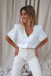 Blouse col V manches brodées portée par une femme, vue de trois-quarts, mains sur les hanches, pantalon crème