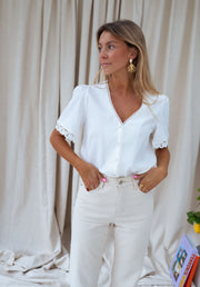 Blouse col V manches brodées portée par une femme, silhouette entière, rideau beige, mains dans les poches