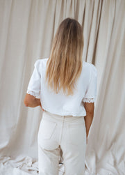 Blouse col V manches brodées portée par une femme, vue de dos, rideau beige, cheveux
