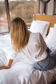 Chemise fluide à cœurs brodés portée par une femme, vue de dos, sur un lit, cheveux longs