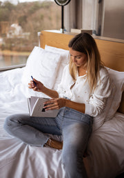 Chemise fluide à cœurs brodés portée par une femme, assise, sur un lit, carnet, jean