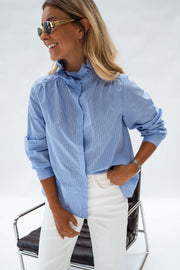 Chemise opaque à col victorien bleue portée par une femme, vue de face, fauteuil en cuir, lunettes de soleil