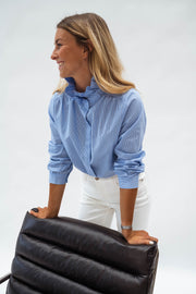 Chemise opaque à col victorien bleue portée par une femme, vue de trois-quarts, fauteuil noir, pantalon blanc