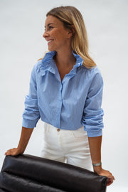 Chemise opaque à col victorien bleue portée par une femme, vue de trois-quarts, fauteuil noir, cheveux détachés