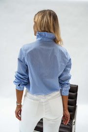Chemise opaque à col victorien bleue portée par une femme, vue de dos, fauteuil noir, pantalon blanc