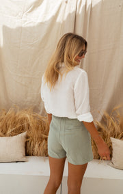 Chemise à col et boutons portée par une femme, vue de profil, rideau beige, short vert sauge
