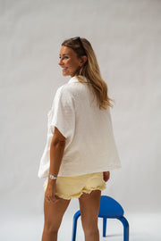 Chemise fluide en plumetis portée par une femme, vue de profil, short jaune, tabouret bleu