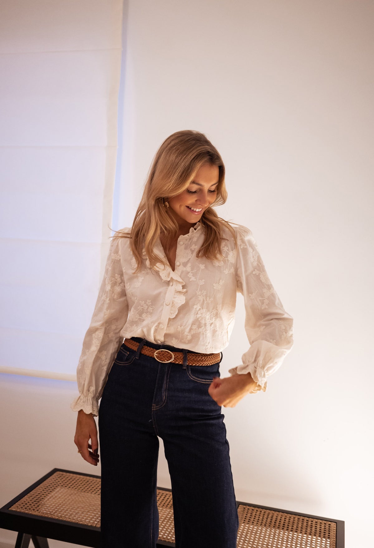Chemise brodée à volants portée par une femme, silhouette entière, pantalon foncé, ceinture marron, banc en cannage