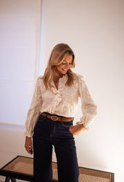 Chemise brodée à volants portée par une femme, silhouette entière, pantalon foncé, ceinture marron, banc en cannage