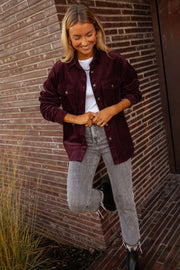 Chemise en velours ample bordeaux portée par une femme, vue de face, mur en briques, jean gris