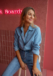 Chemise en denim fluide portée par une femme, vue de trois-quarts, sourire, jean, mur rose