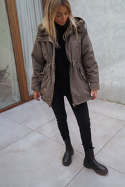Manteau imperméable à capuche fourrée porté par une femme, silhouette entière, manteau kaki, bottines noires, mur gris