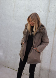 Manteau imperméable à capuche fourrée porté par une femme, vue de trois-quarts, mains dans les poches, mur gris