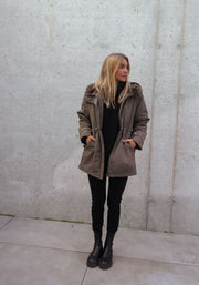 Manteau imperméable à capuche fourrée porté par une femme, silhouette entière, de loin, mur gris