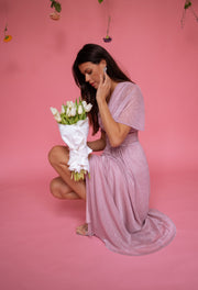 Robe longue à lurex brillant portée par une femme, assise, fond rose, bouquet de fleurs blanches
