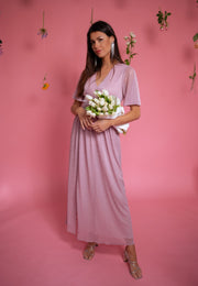 Robe longue à lurex brillant portée par une femme, vue de face, fond rose, bouquet blanc