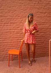 Combishort fluide ouverture buste porté par une femme, vue de face, chaise orange, main sur le nœud