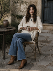 Pull blanc cassé ourlet asymétrique — vue portée assise sur une chaise en bois sur une terrasse, bottes marron