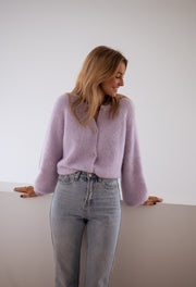Gilet à boutons fleuris brillants porté par une femme, vue de trois-quarts, adossée à un meuble blanc, sourire