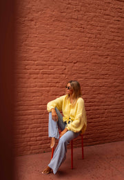 Gilet à liens noués jaune porté par une femme, assise, chaise orange, mur de briques
