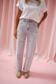 Jeans fleuri taille haute porté par une femme, silhouette entière, rideaux roses, t-shirt blanc