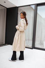 Trench ample ceinturé à poches porté par une femme, vue de dos, bottines noires, baie vitrée