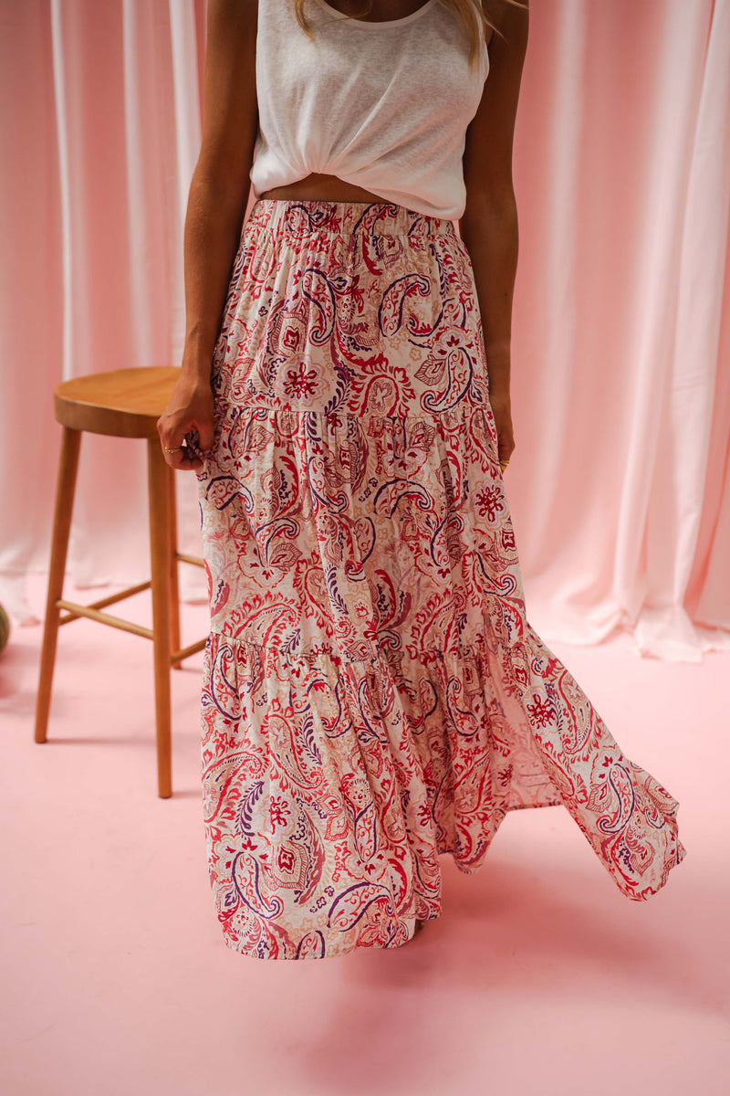 Jupe longue fluide à motifs portée par une femme, en marchant, rideaux roses, tabouret, jupe fleurie