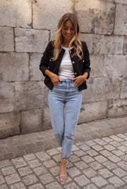 Veste droite à épaulettes portée par une femme, silhouette entière, jean clair, mur en pierre