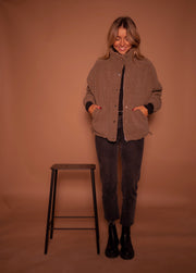 Manteau mouton à col montant porté par une femme, silhouette entière, mains dans les poches, tabouret noir